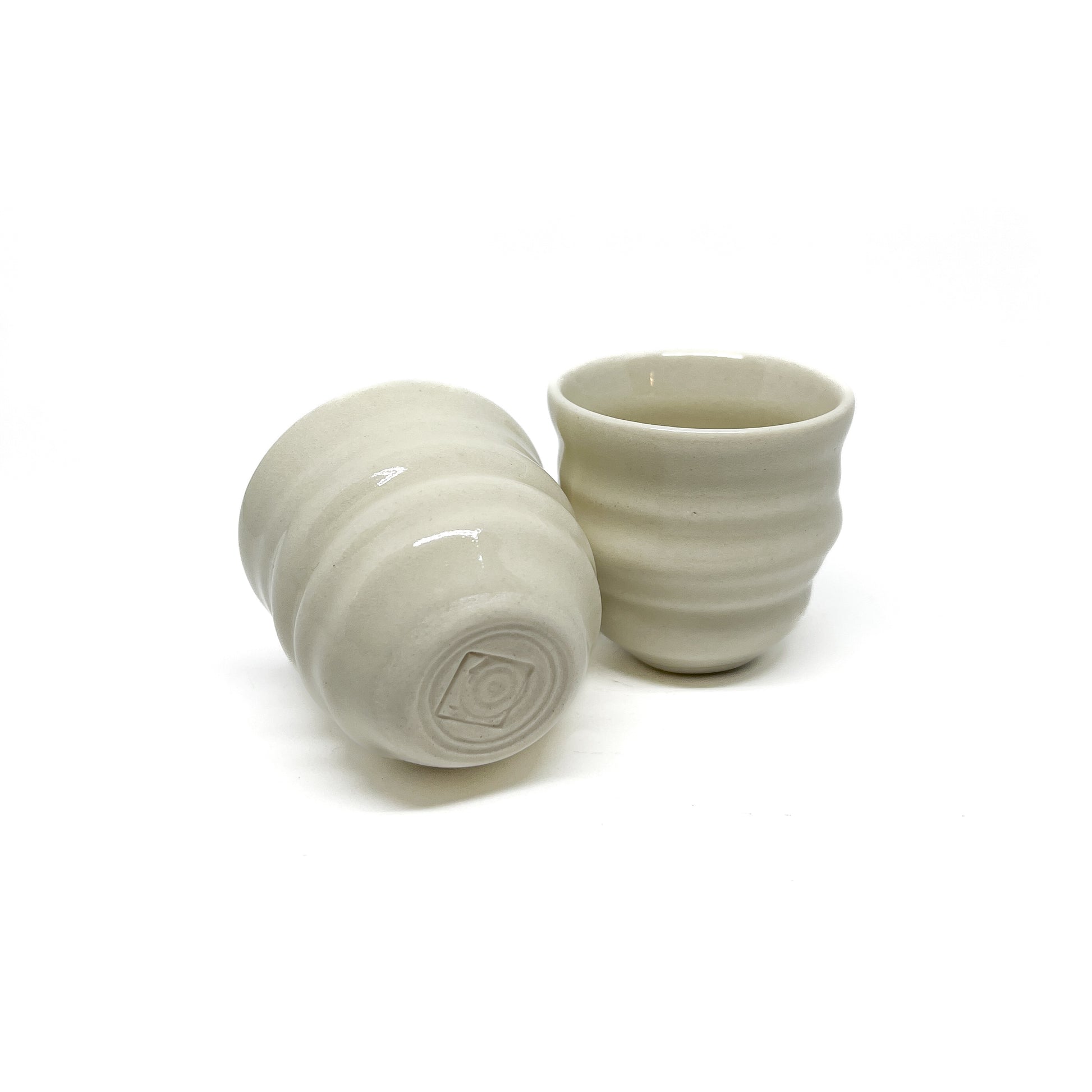 Two small ceramic Yunomi cups with subtle horizontal ridges. One is upright, the other is tilted showing a circular base with a geometric mark.