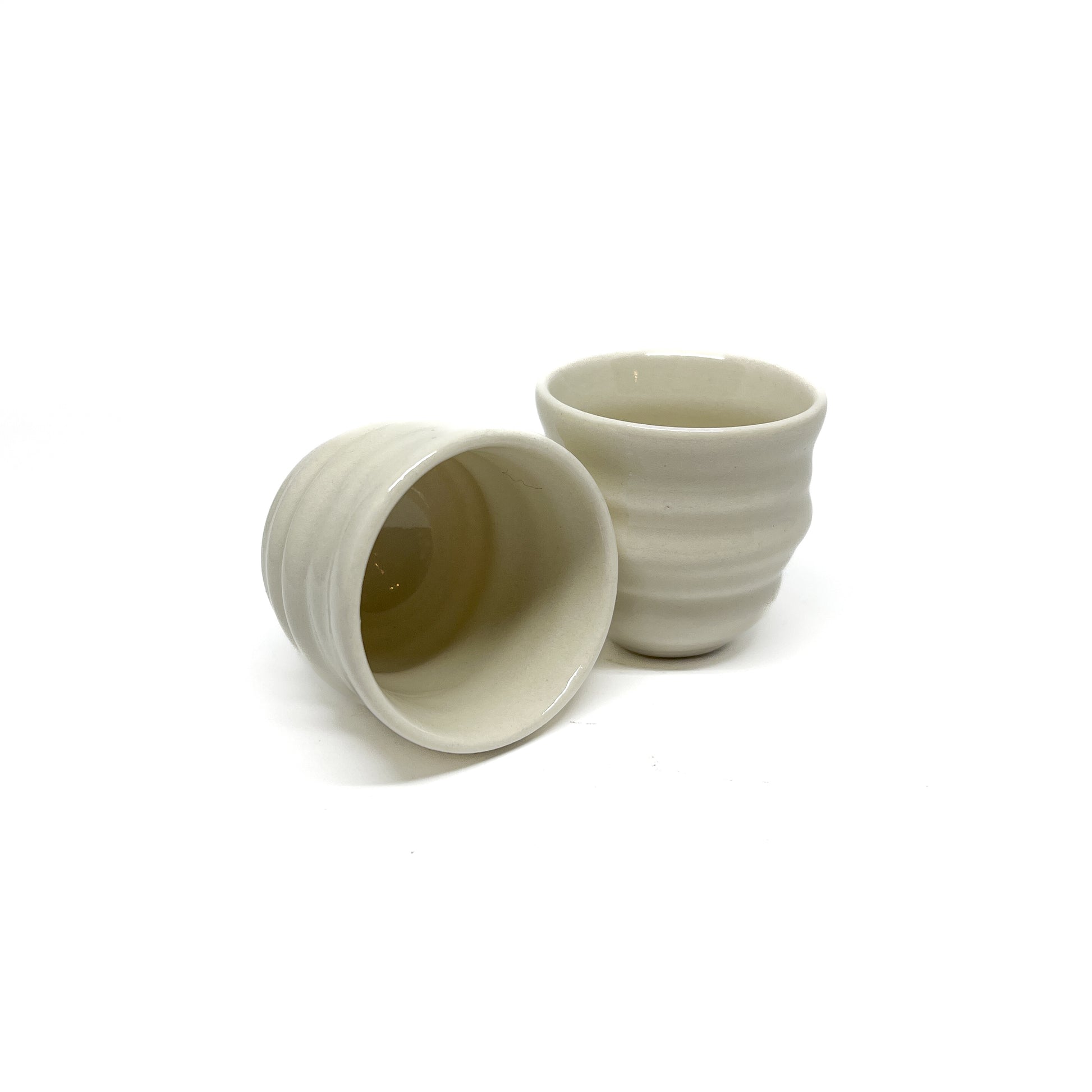 Two small ceramic Yunomi cups with subtle horizontal ridges. One is tilted, showing its hollow interior, next to an upright cup.