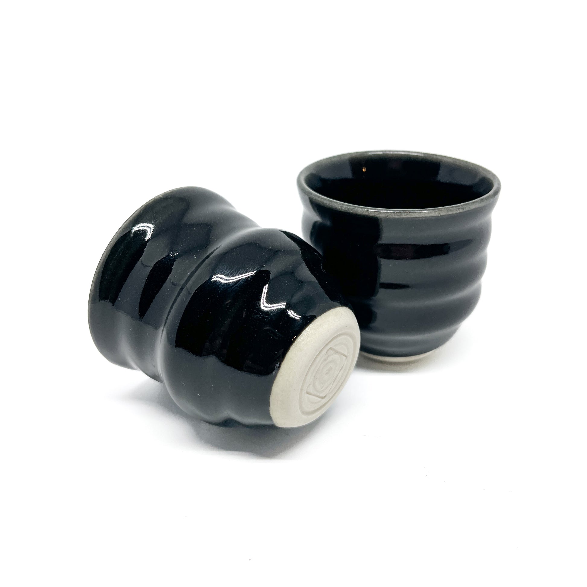 Two small, dark Mino ware sake cups with horizontal ridges. One is upright, the other is tilted showing a light-toned base with concentric circles.