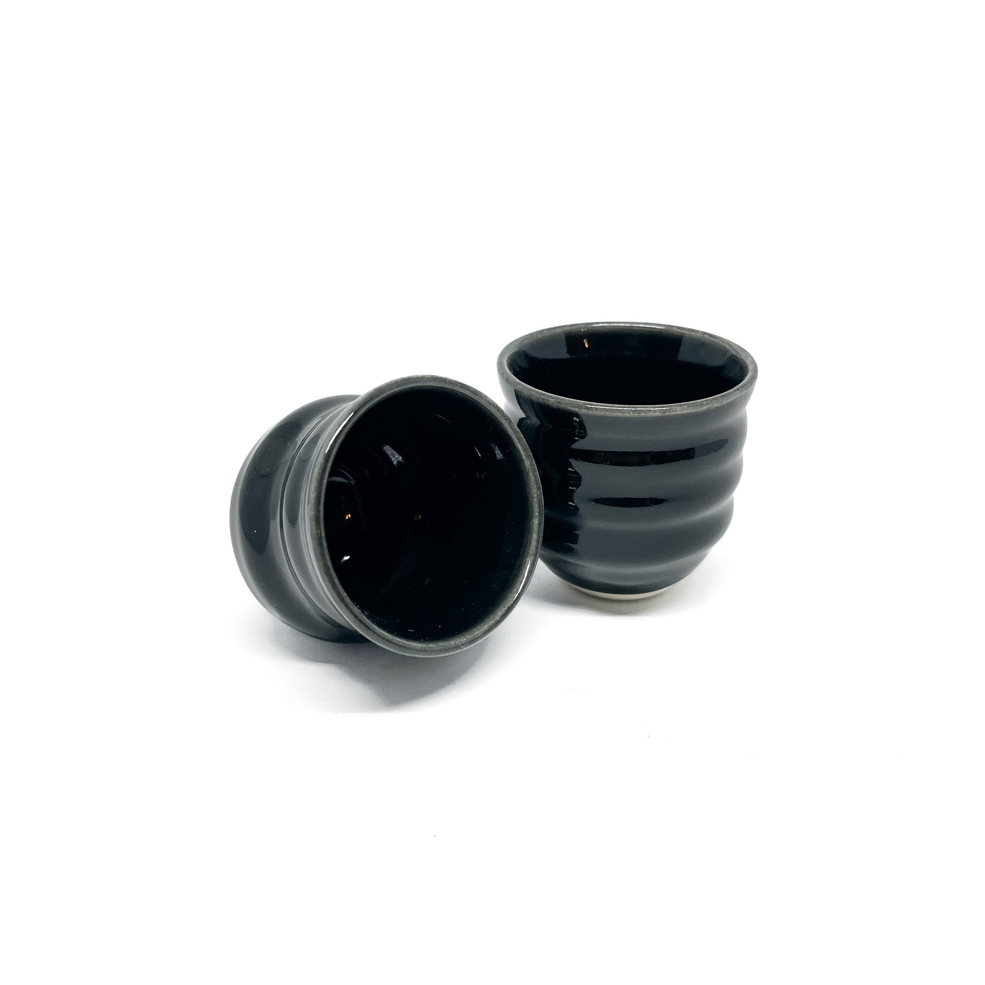 Two small, dark Mino ware sake cups with horizontal ridges. One is tilted, showing its dark interior, next to an upright cup.