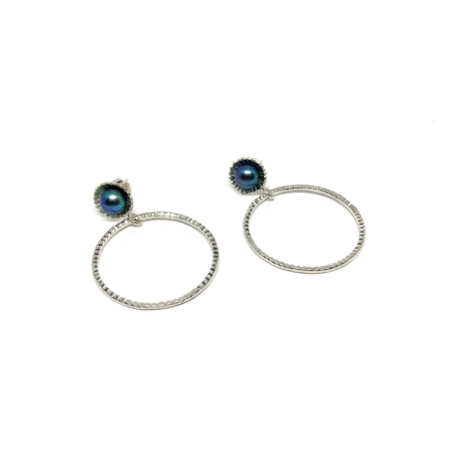 A pair of silver hoop earrings, each adorned with a dark, shimmering Taku Poe pearl set above a textured circular hoop.