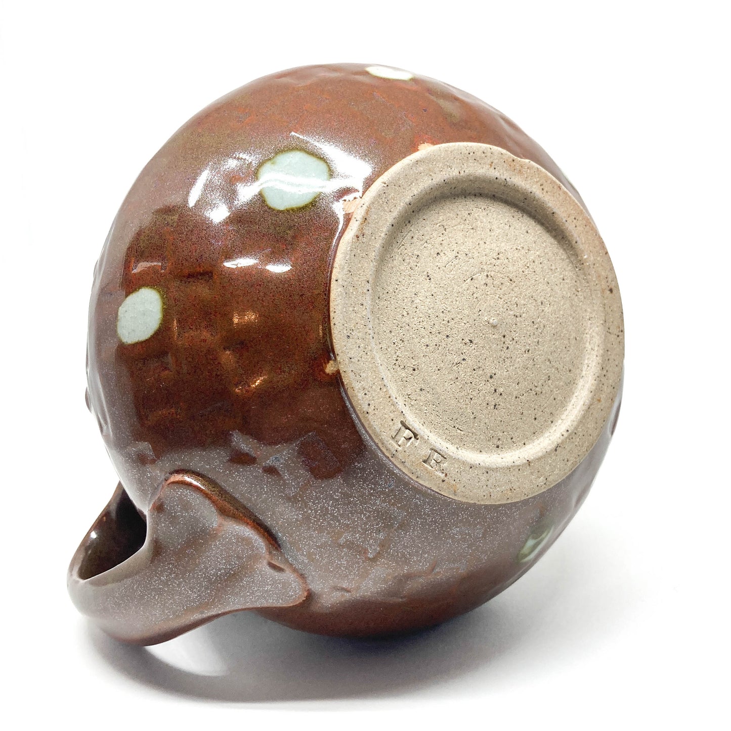 Bottom view of a round, brown teapot with light polka dots and an unglazed base showing maker's marks. Handle visible.