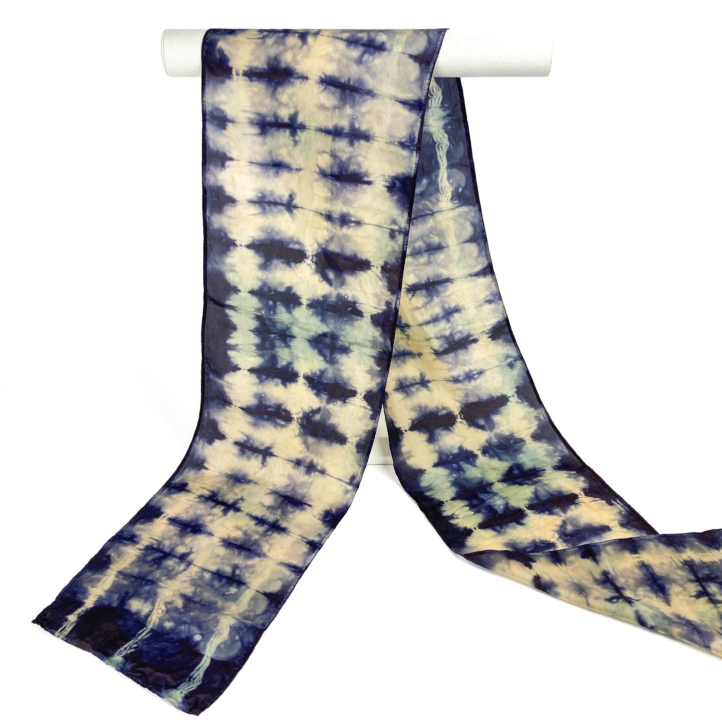 An Indigo and Myrobalan silk scarf with a textured, resist shibori pattern hangs from a white cylindrical bar.