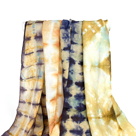Five silk scarves with unique shibori tie-dye patterns in various color palettes hang from a white bar.