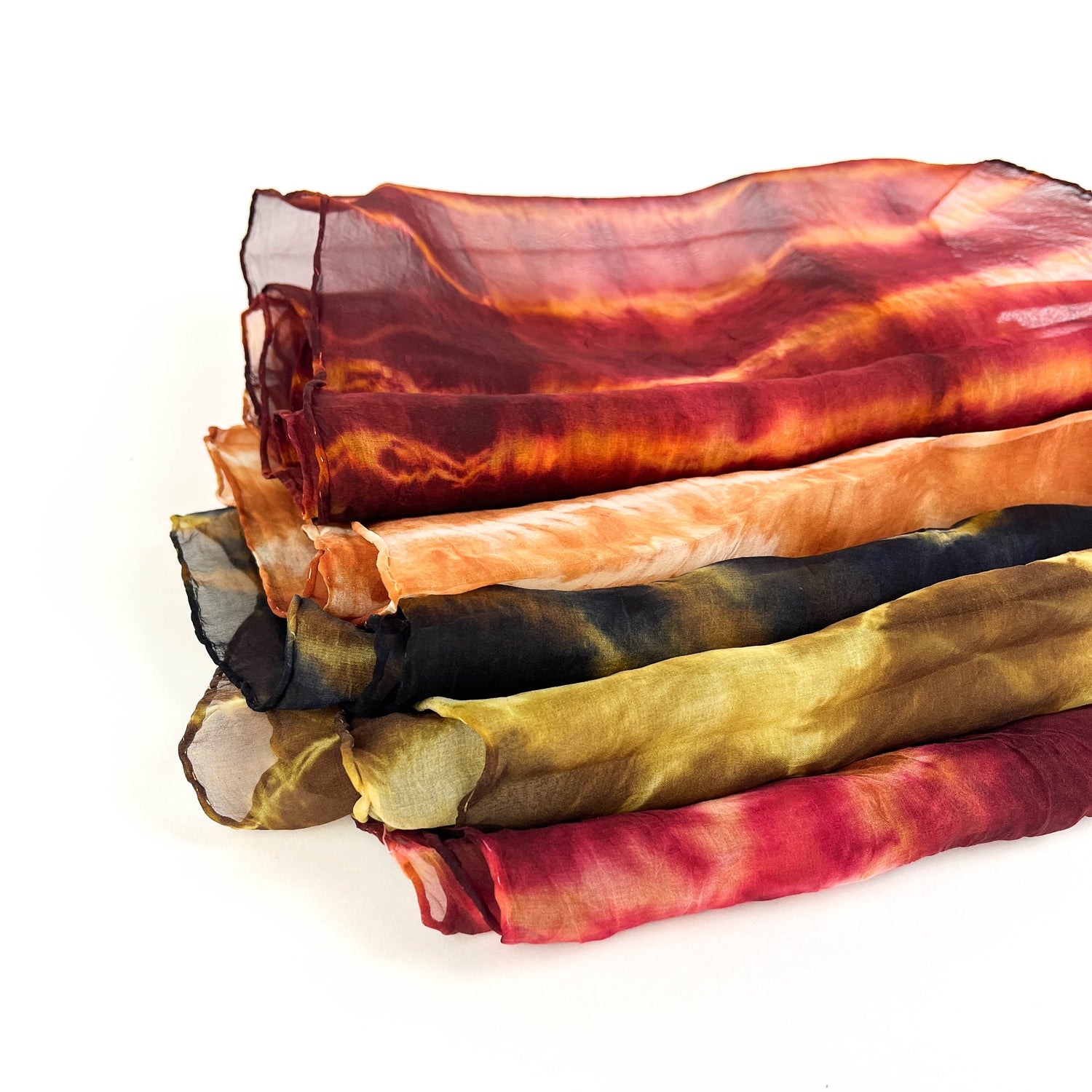 A neatly stacked pile of six sheer silk scarves showcases a variety of vibrant, blended tie-dye patterns and textures.