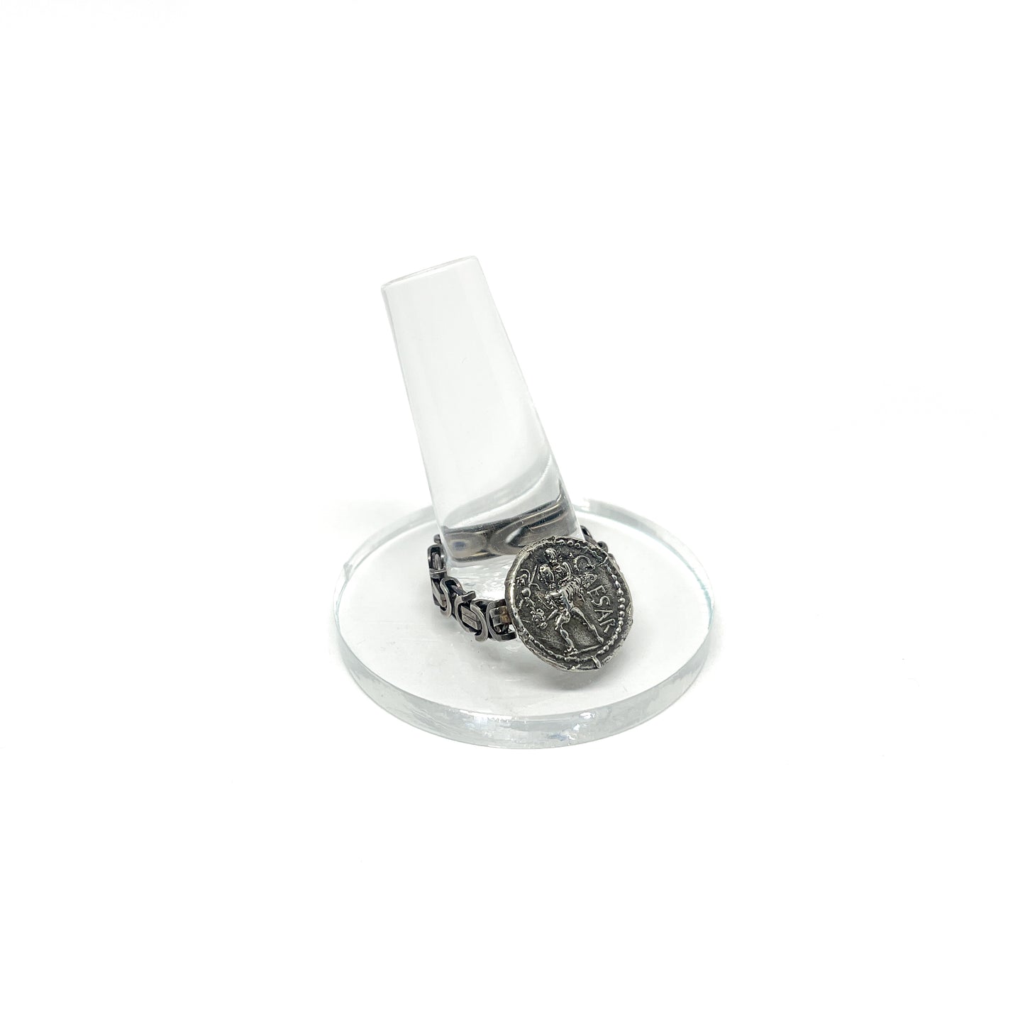 A silver ring with a chain band and an antique coin featuring ".CAESAR" and a figure, displayed on a clear ring stand.