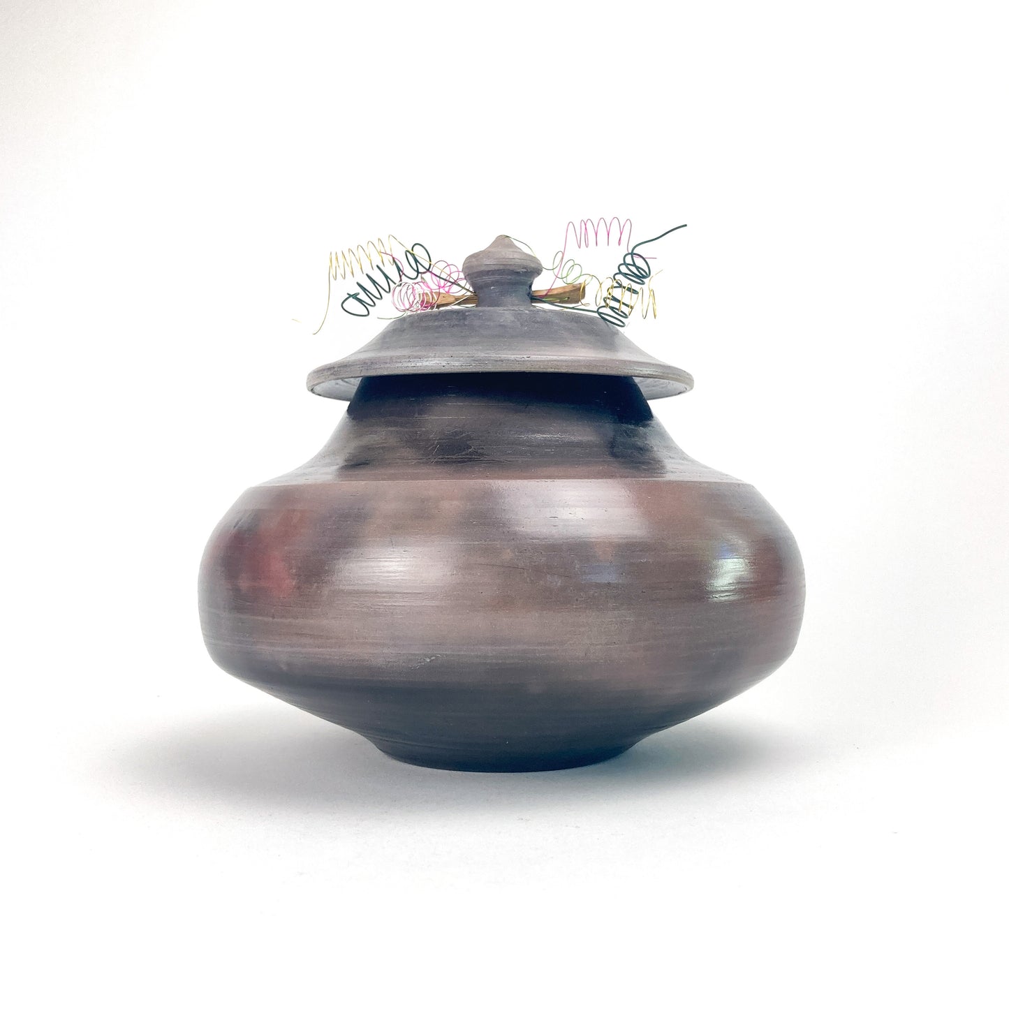 Lidded ceramic jar with smooth burnished surface, topped with decorative multicolored wire elements.