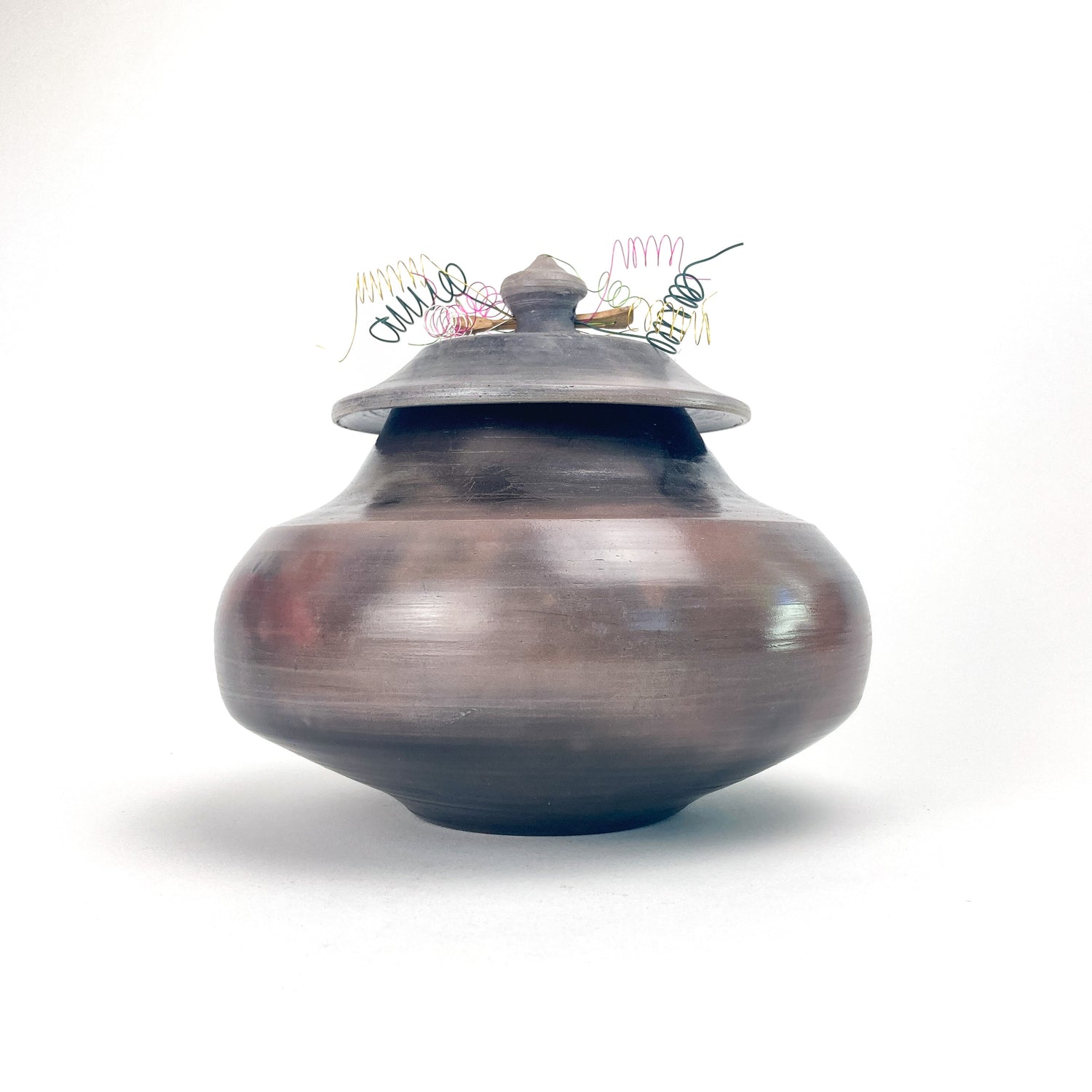 Lidded ceramic jar with smooth burnished surface, topped with decorative multicolored wire elements.