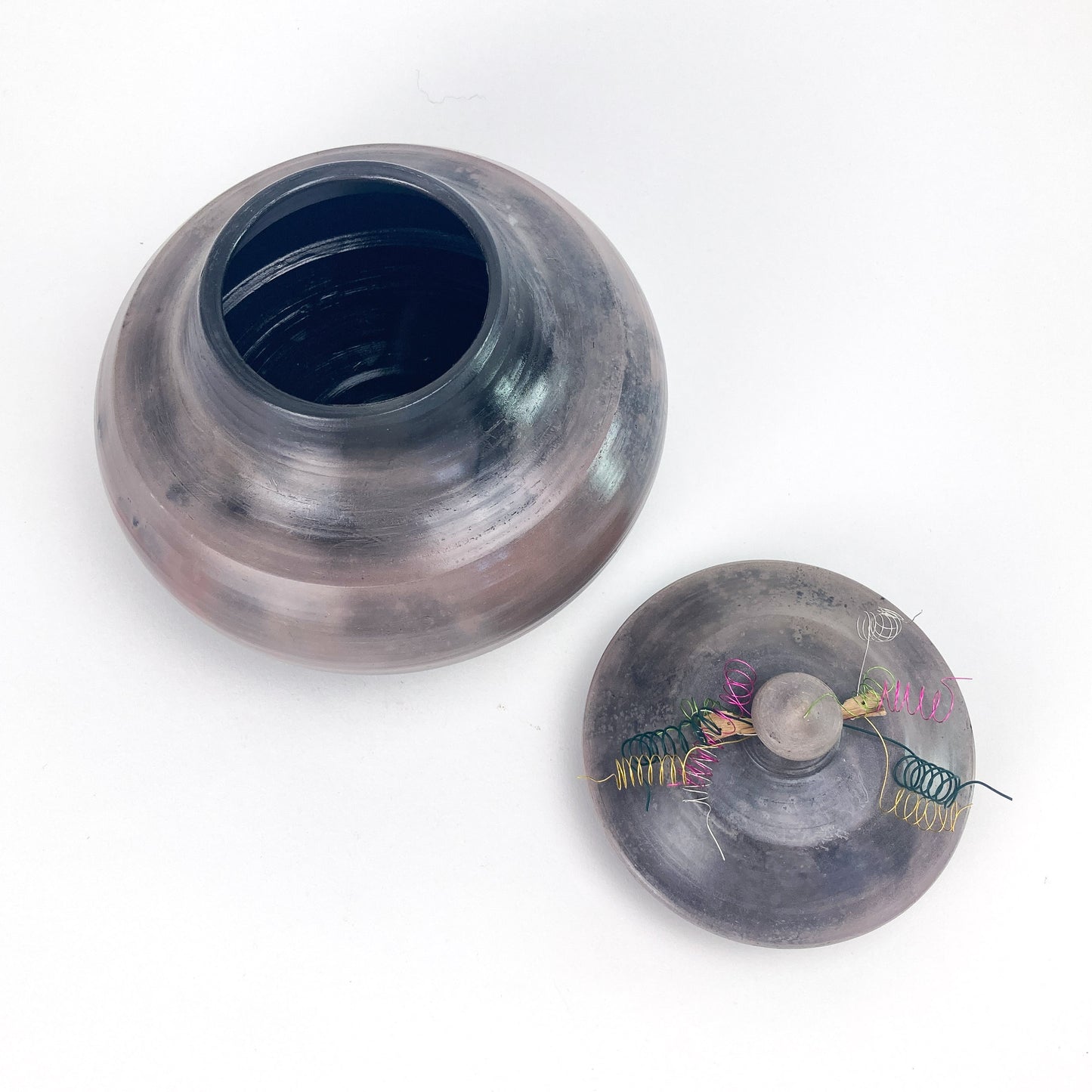 An open, textured jar sits beside its lid, which features colorful wire spirals, a central stone knob, and a visible coil spring.