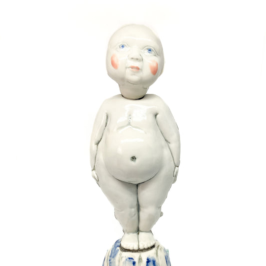 A white ceramic figurine of a child with blue eyes and rosy cheeks stands on a decorated base.