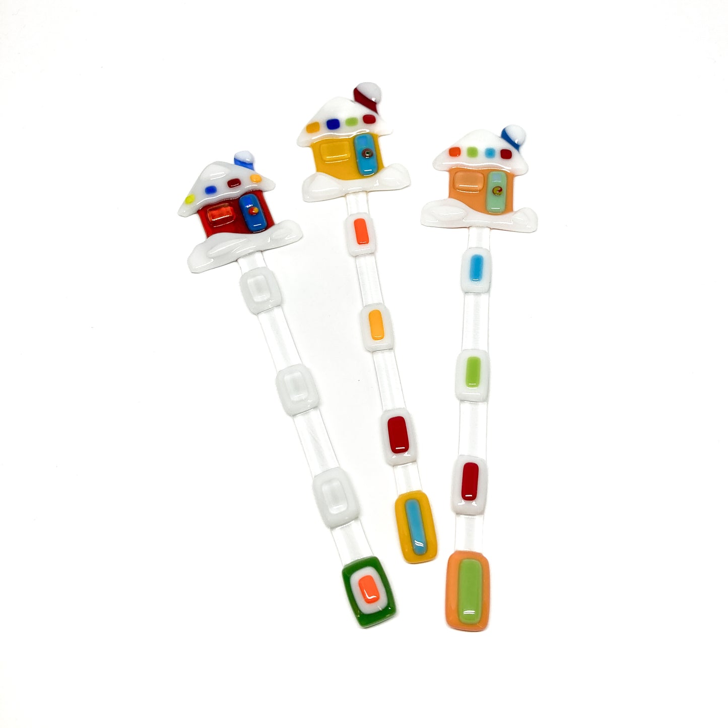 Three glass stir sticks shaped like houses, decorated with colorful glass pieces.