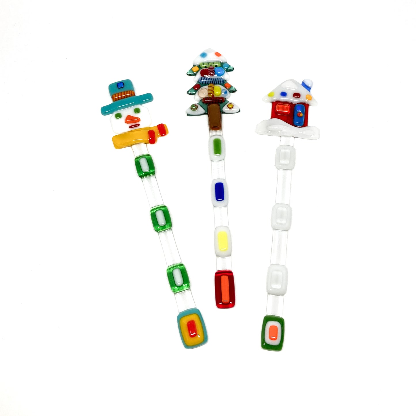 Three glass stir sticks shaped like a snowman, tree, and house, decorated with colorful glass pieces.
