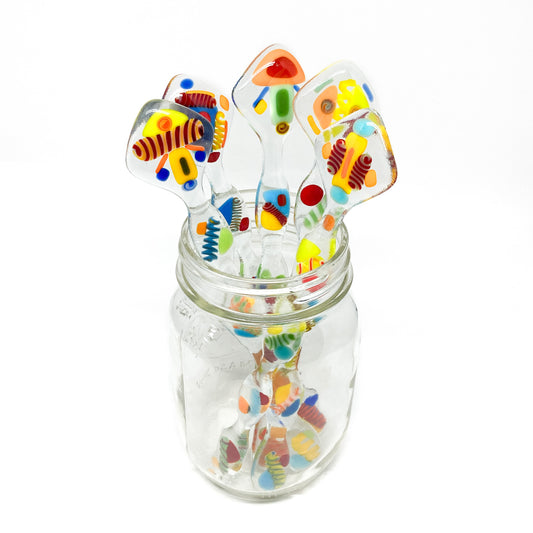 Five glass stir sticks with colorful abstract designs, arranged in a clear glass jar.