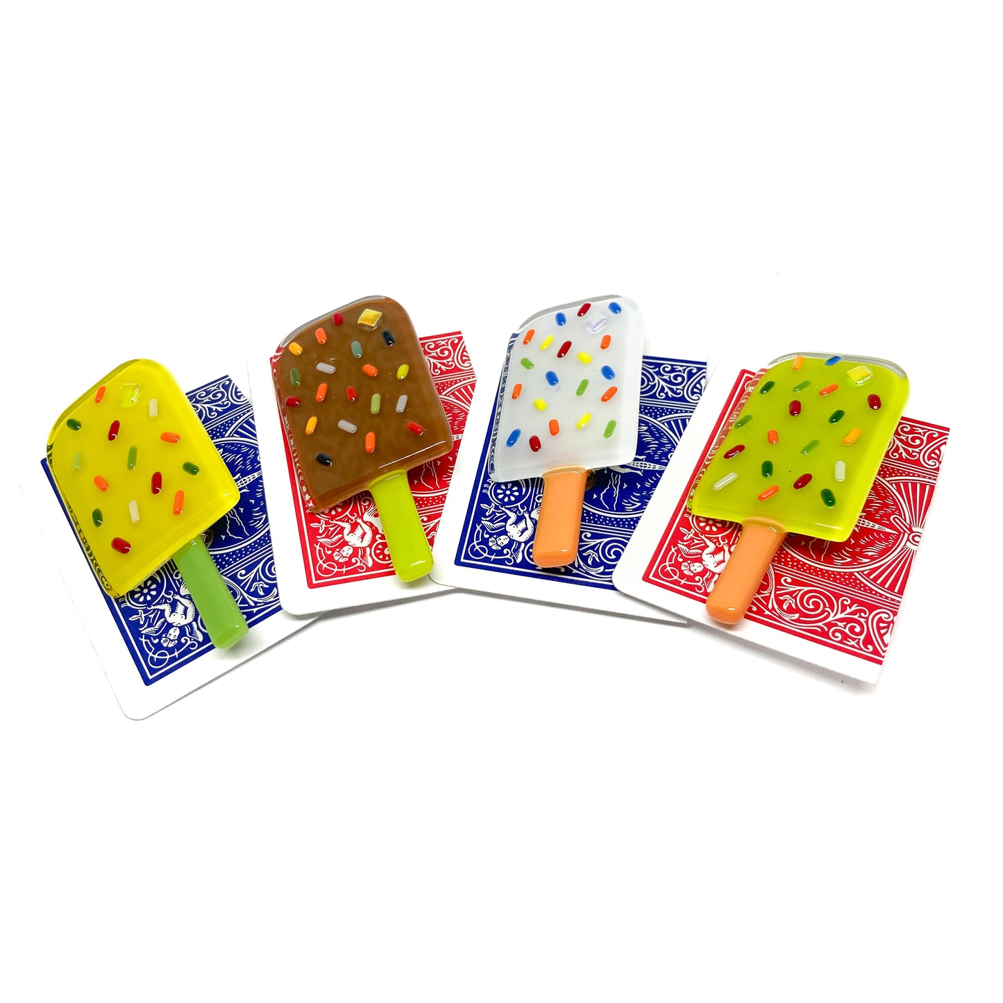 Four glass pins shaped like popsicles in different colors, each placed on a playing card.