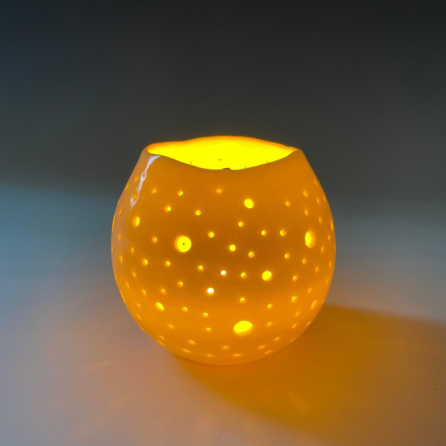 A round ceramic tealight holder with small holes, glowing from the light of a candle inside.