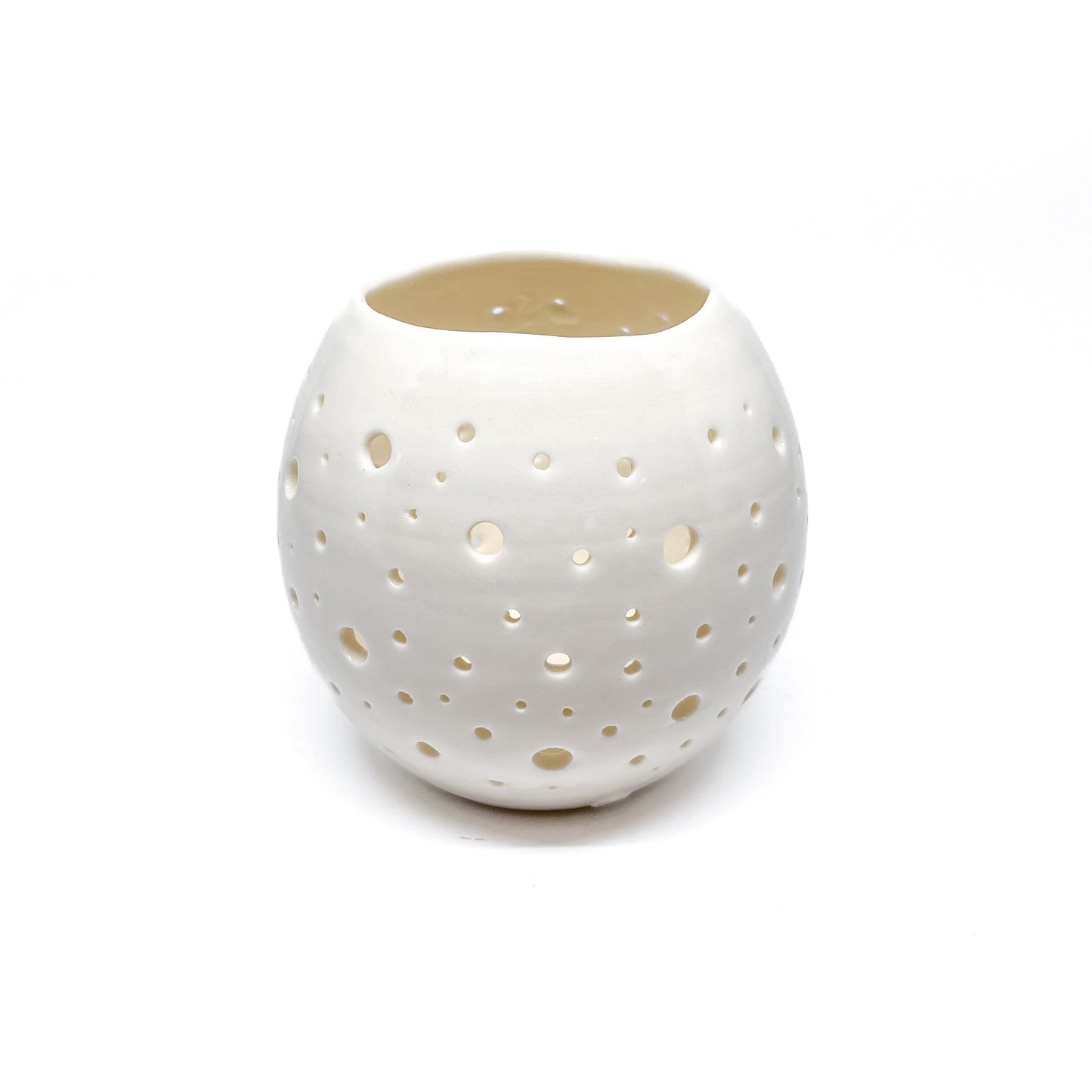 A round, white ceramic tealight holder with small holes cut out around its surface, seen from a slightly different angle.