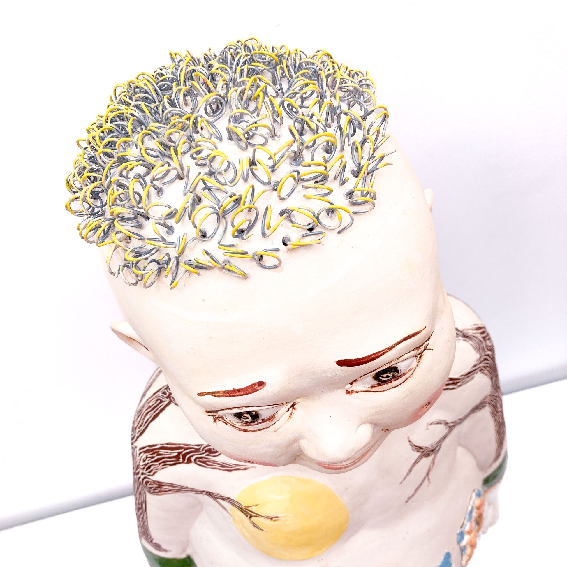 A baby sculpture with a bald head, painted eyes, and metal rings for hair, viewed from above.