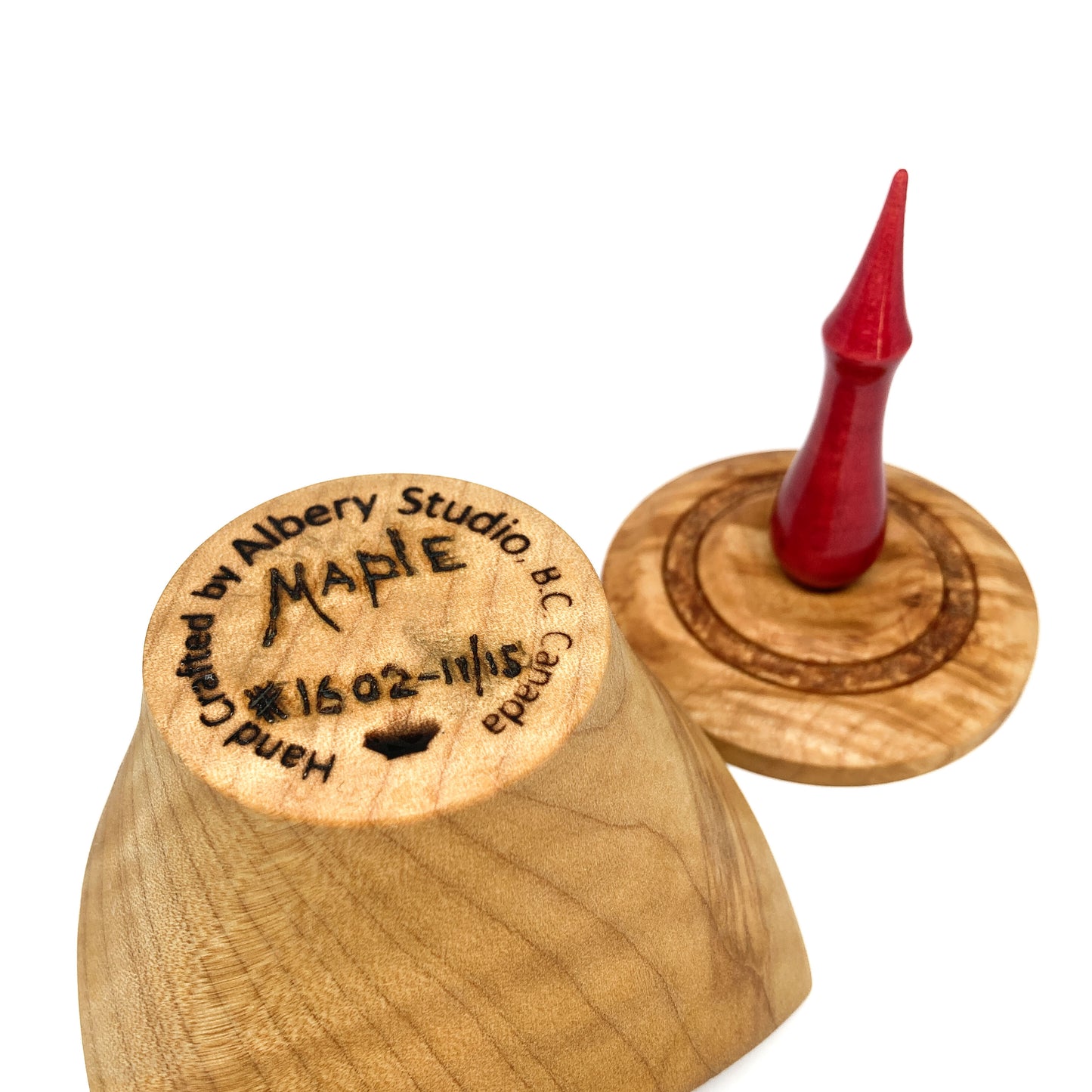 A turned wooden box lid with a pointed red finial, showing the maker's mark on the bottom.