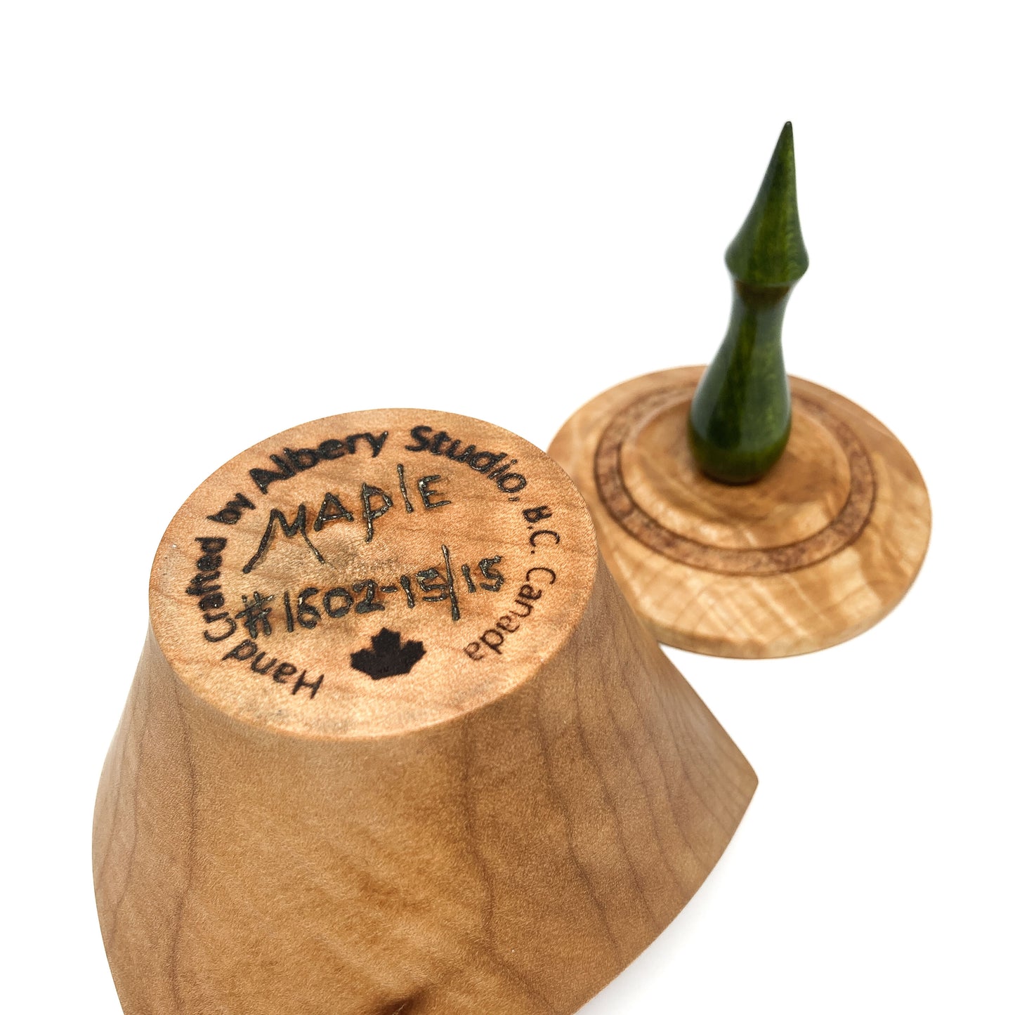 A turned wooden box lid with a pointed green finial, showing the maker's mark on the bottom.