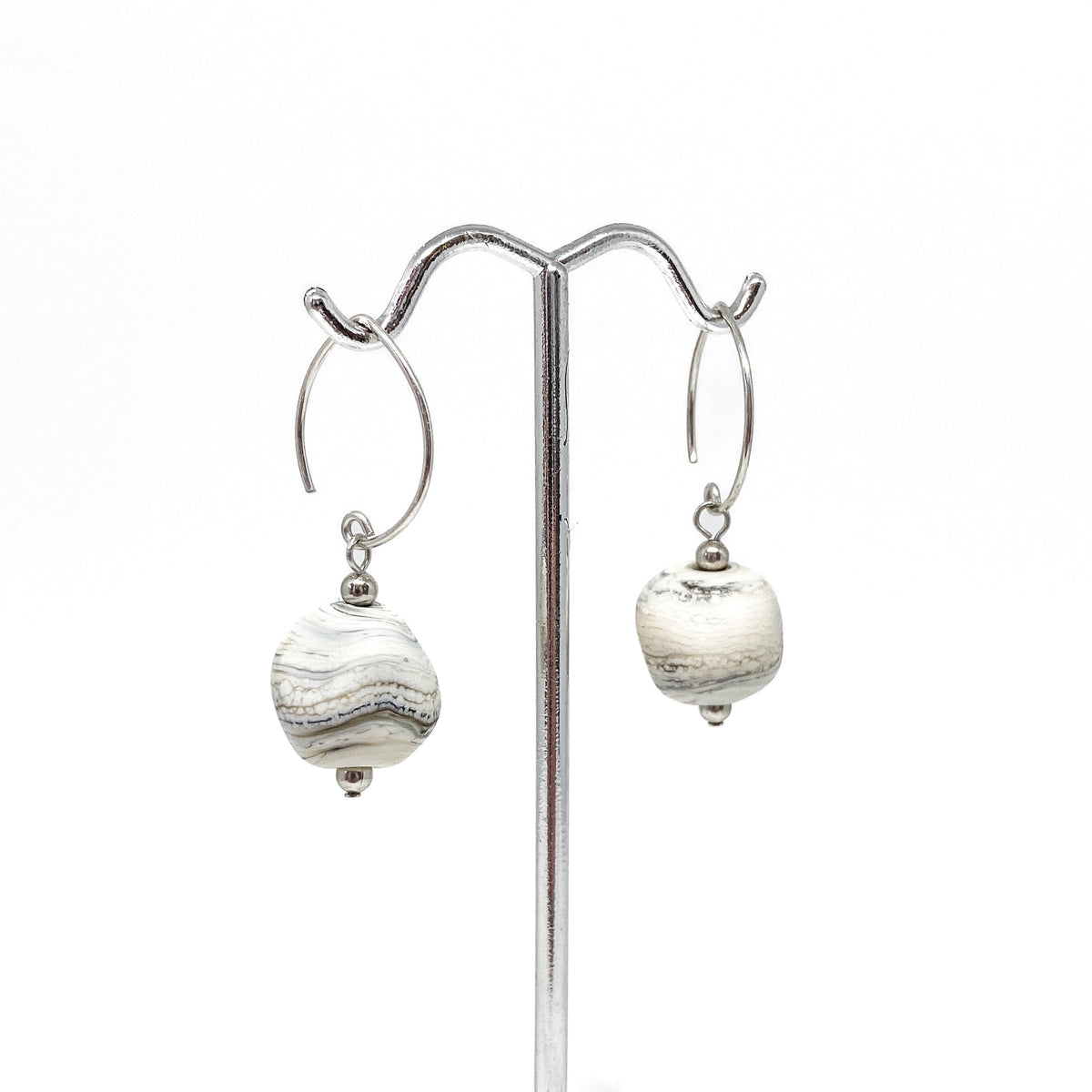 A pair of earrings with gray and white swirl glass beads hanging from silver hoops on a metal stand.