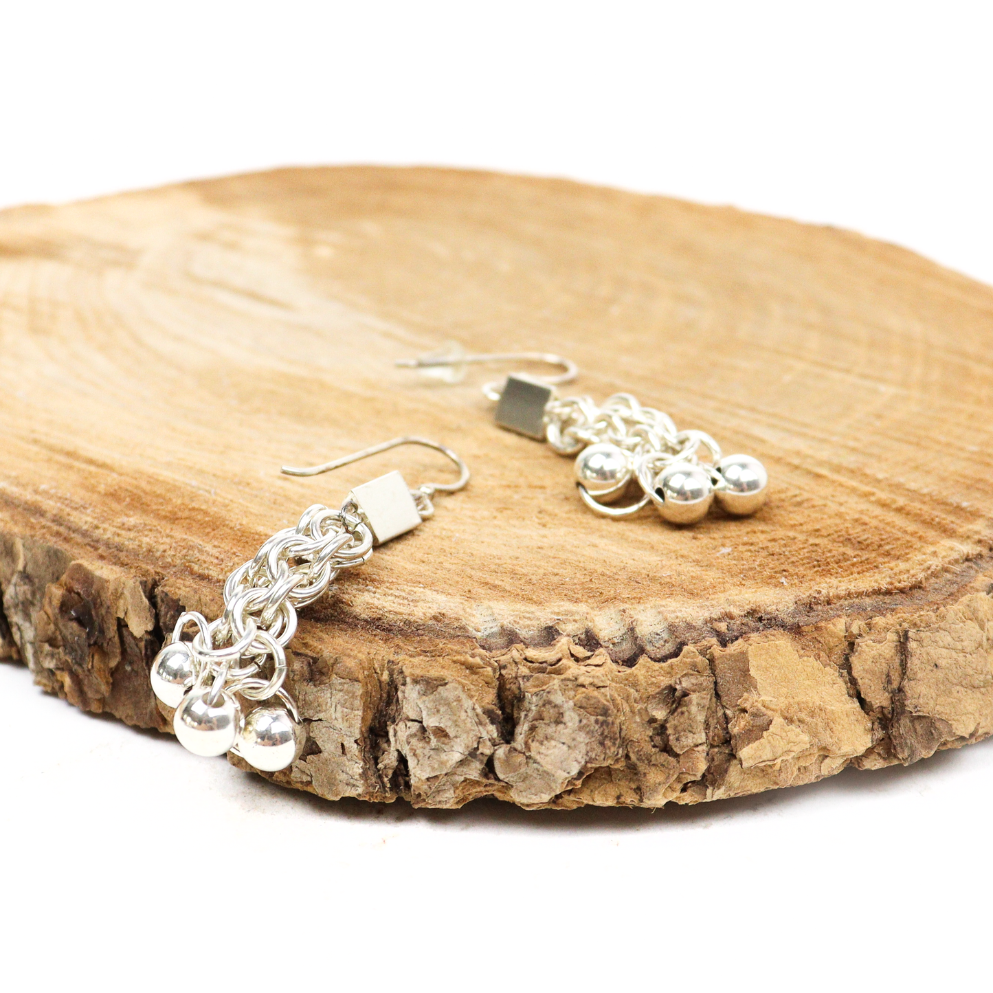 Chainmail dangle earrings resting on a textured wooden surface, showcasing linked rings and ball details.