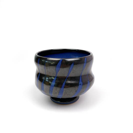A dark ceramic tea bowl with blue diagonal stripes, resting on a small pedestal.
