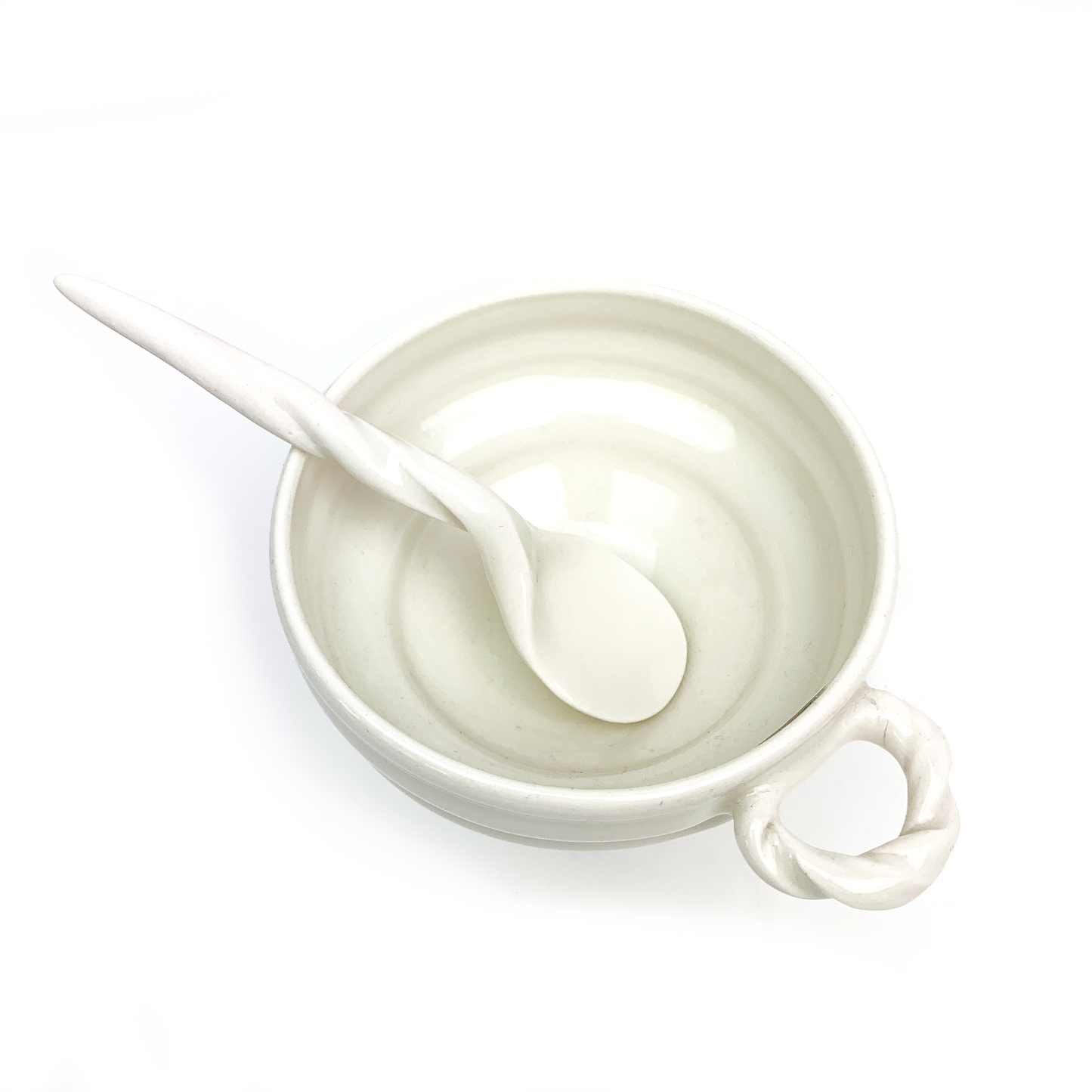 Top-down view of a round ceramic bowl with a twisted handle and a matching spoon resting inside.