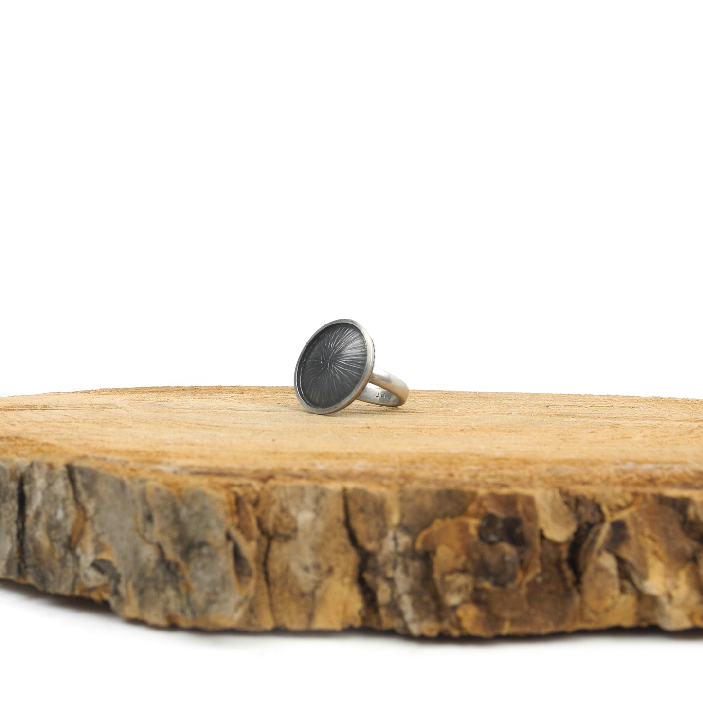 Silver ring with a large, round, dark textured face featuring a radiating design, resting on a textured surface.