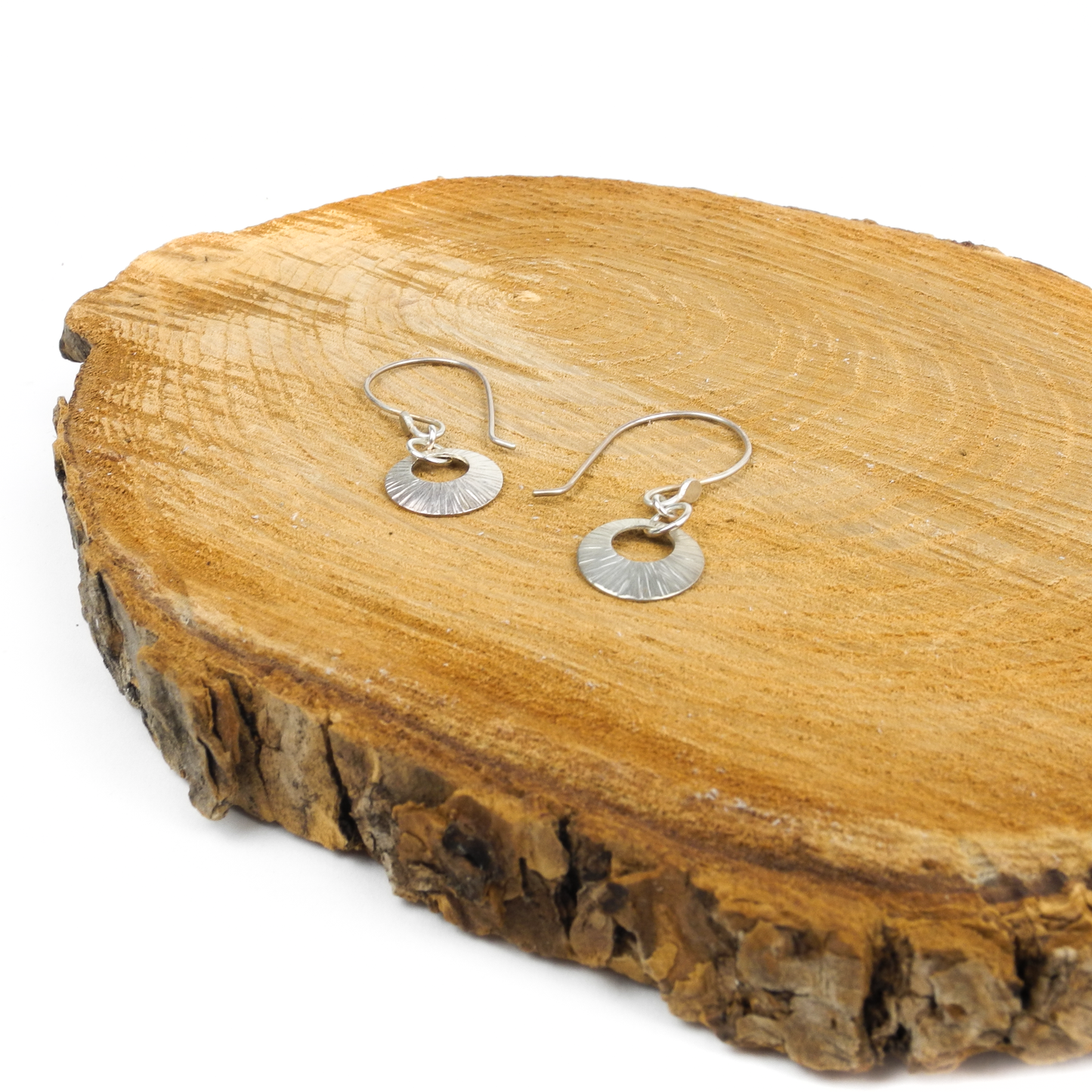 Two silver earrings with radiating lines and a center cutout sit on a textured wooden surface.