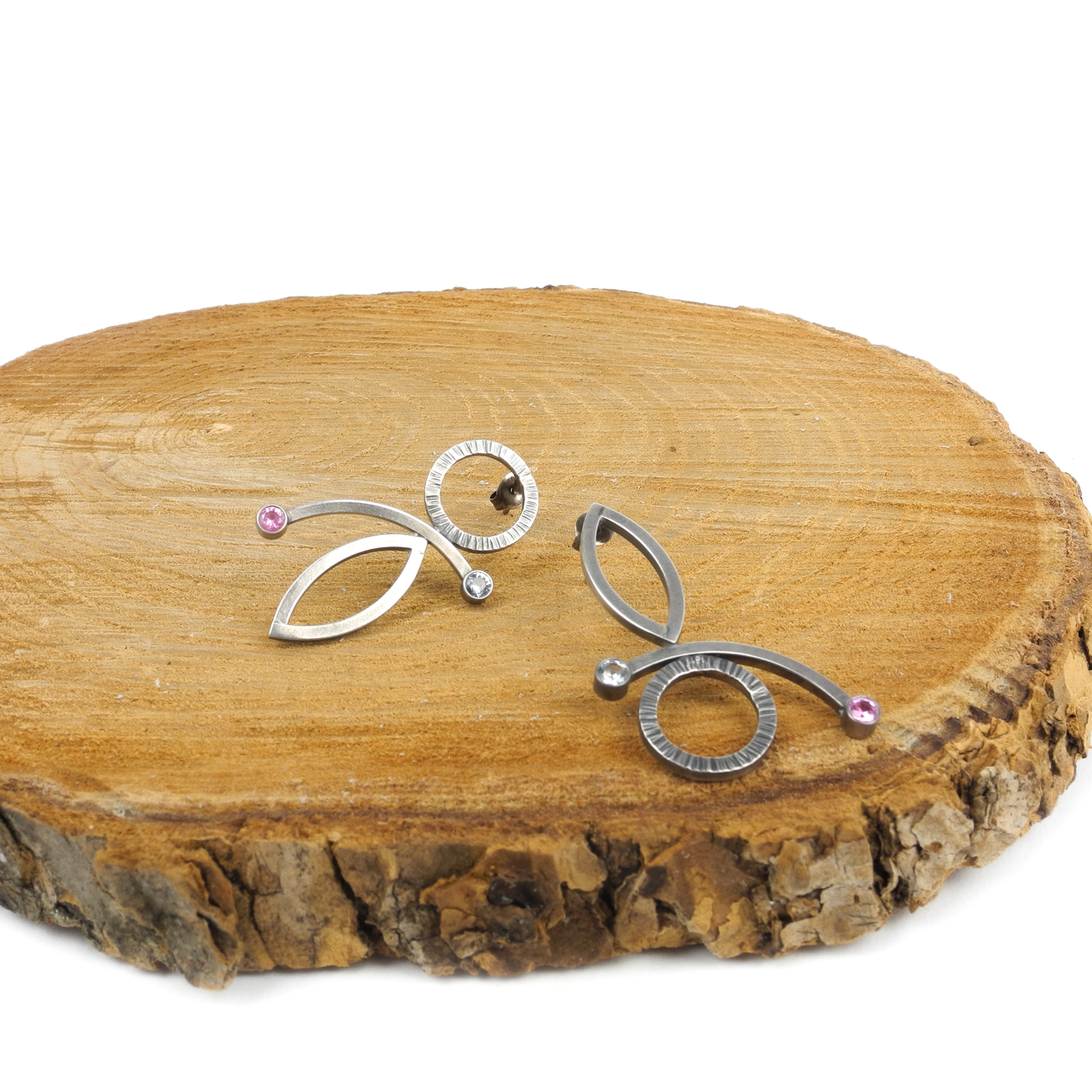 Two abstract metallic earrings with circles, ovals, curves, and small pink and clear gems rest on a wooden surface.