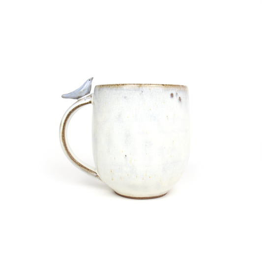 Handmade pottery mug from a pottery shop, featuring a small, sculpted bird perched on the handle. The mug has a textured, off-white glaze.