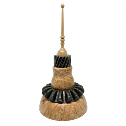 Stacked mixed media spire sculpture with wood, black ridged elements, and a slender, topped spire.