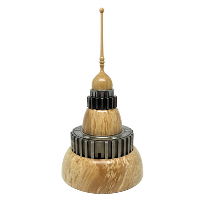 Stacked wood spire sculpture by David Adolph with multiple dark, cog-like rings and a tall, slender, topped spire.