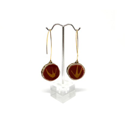 Two round earrings with red and gold bird-shaped designs, displayed on a silver earring stand.