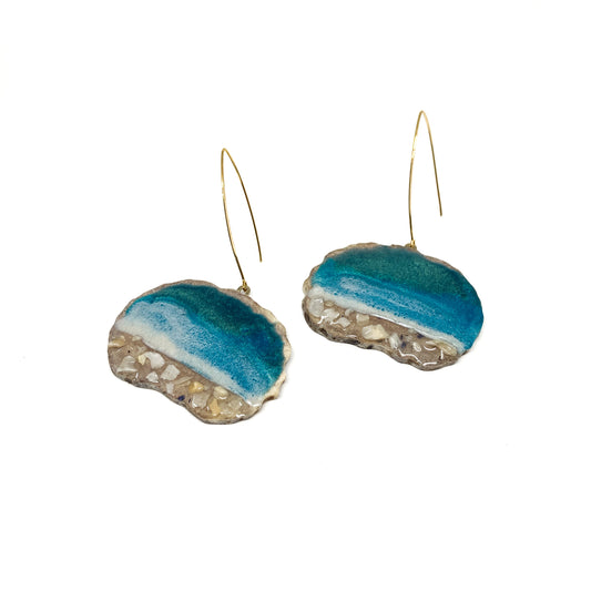 Two round earrings with blue resin and sand-like textures, featuring gold hooks. Ocean-inspired design.