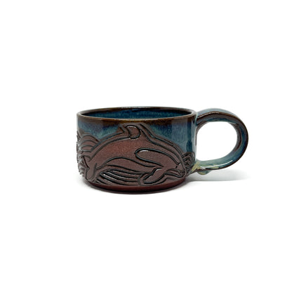 Ceramic mug with a carved whale and wave design in brown against a glossy blue-green glaze and a rounded handle.