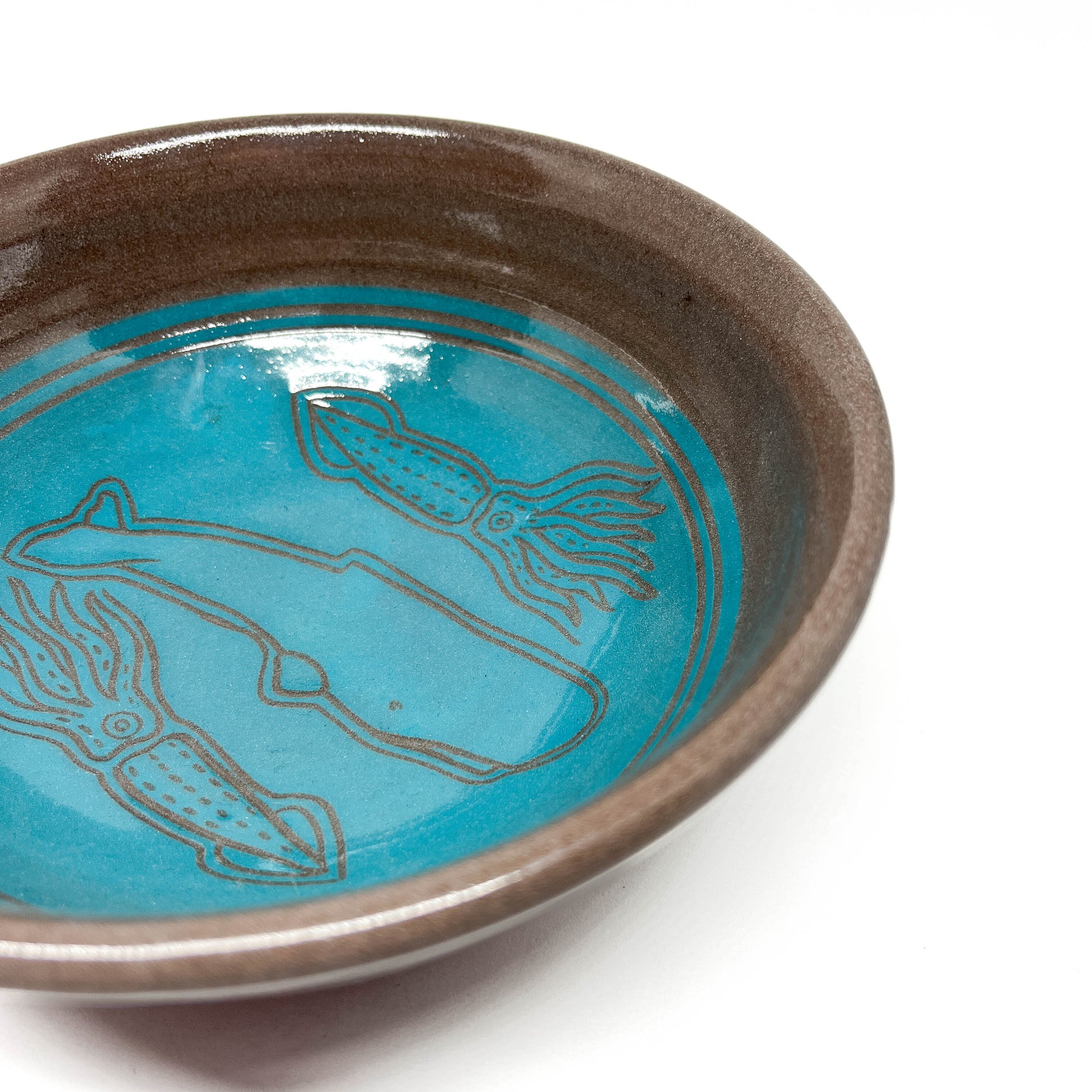 Angled view of a ceramic bowl with etched designs of a whale and two squid in a circular underwater motif.