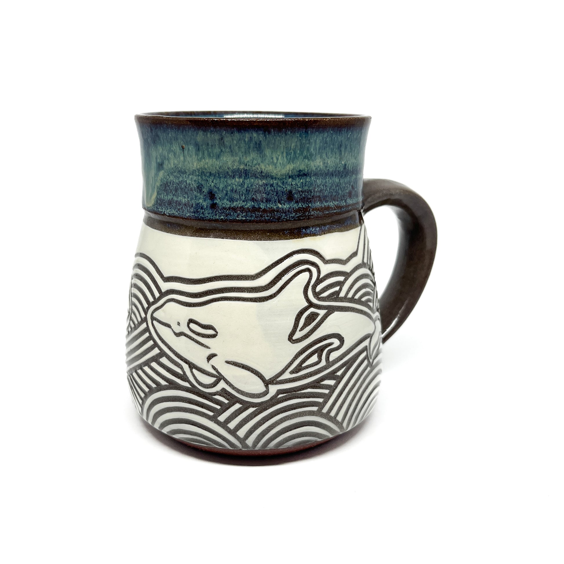 Back view of a carved ceramic mug with two whales diving into stylized waves beneath a glazed rim.