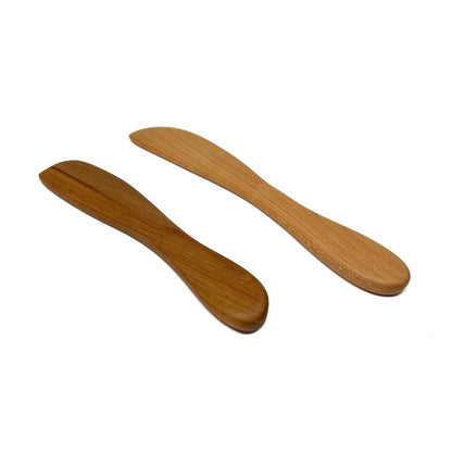 Two wooden spread knives in varying shades, angled on a white surface.