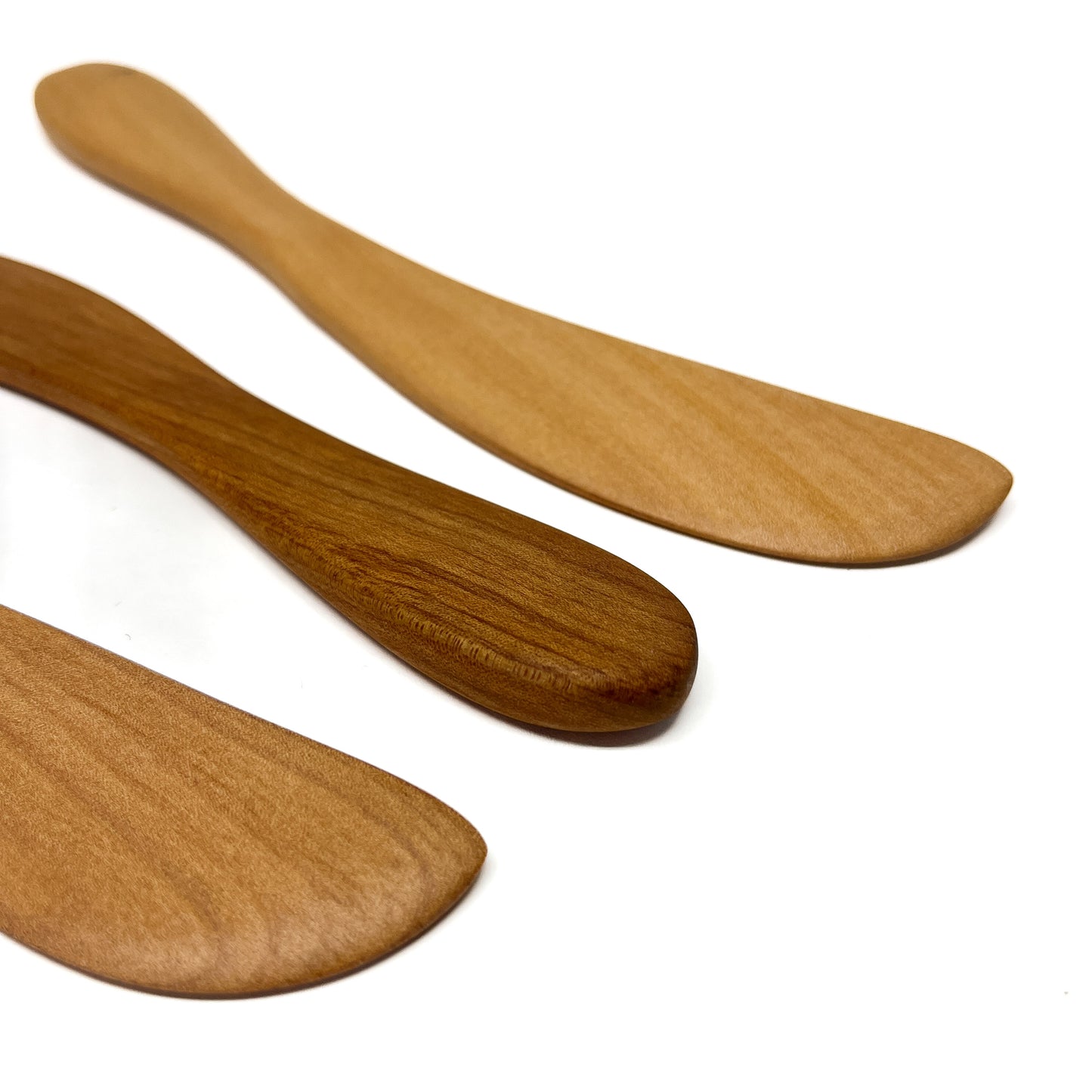 Three wooden spread knives in varying shades, angled on a white surface.