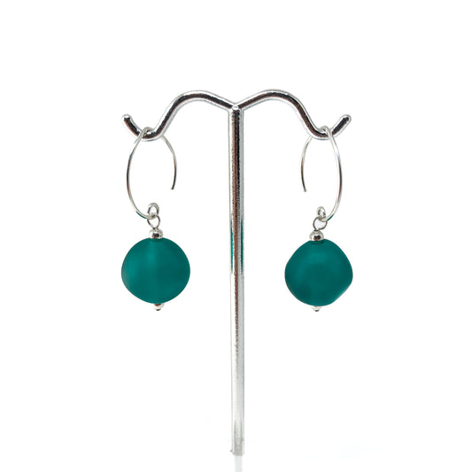 A pair of earrings with teal glass beads hanging from silver hoops on a metal stand.