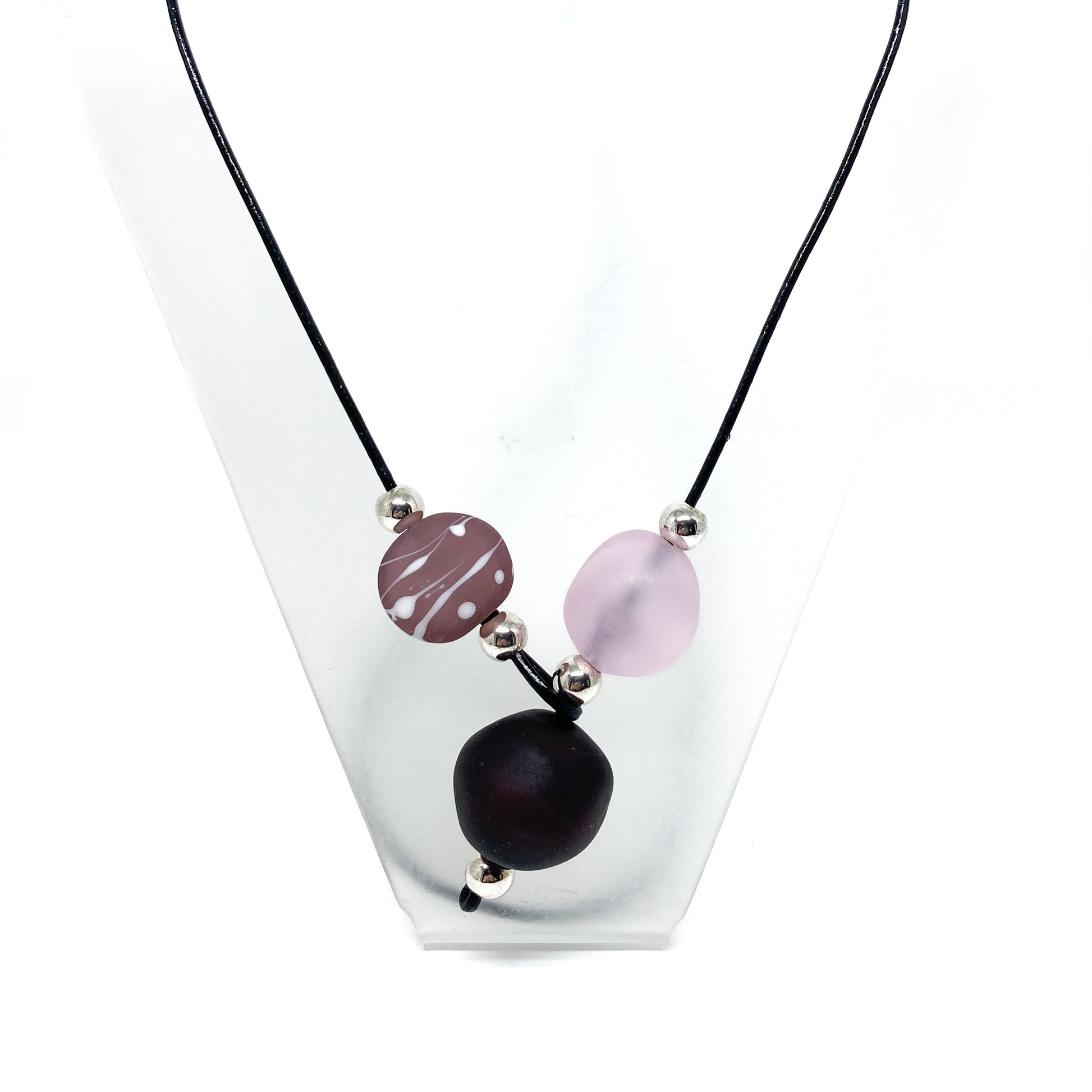 Black cord necklace with purple patterned, dark, and light pink beads, and silver accents, displayed on a stand.