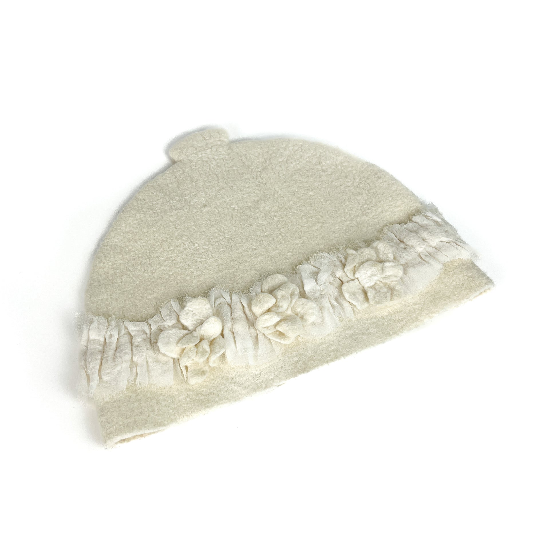 Angled view of an off-white felted teapot cozie with textured floral accents and a fringed edge.
