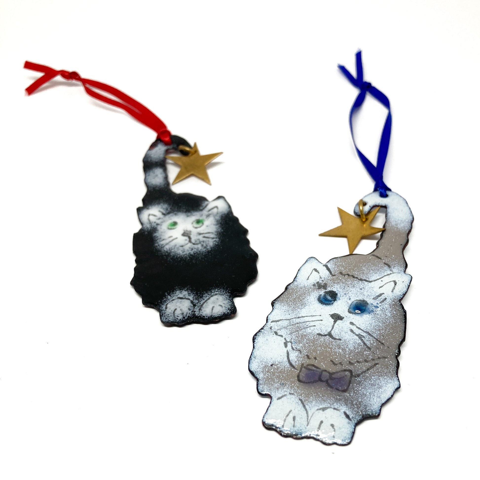 Two cat Christmas ornaments: one black/white with a red ribbon, one gray/white with a blue ribbon, each holding a gold star.