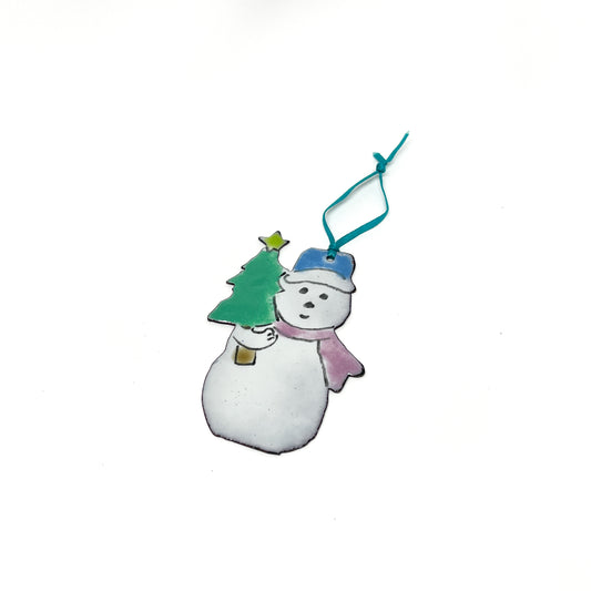 A smiling snowman ornament with a blue hat and pink scarf holds a green Christmas tree with a yellow star. A teal ribbon is attached for hanging.