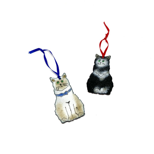 Two sitting cat ornaments: one white with a blue collar, the other black and white with a red ribbon.