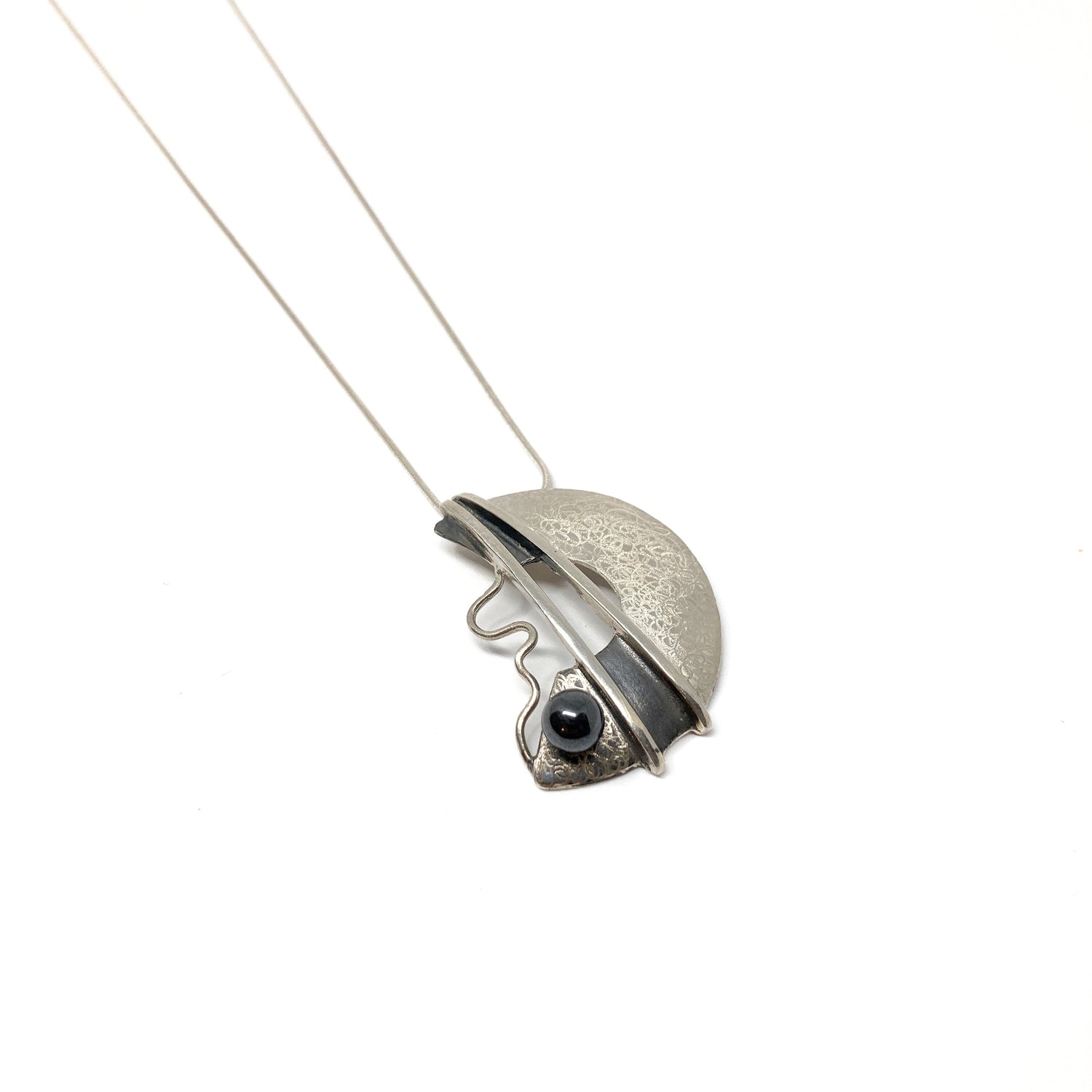 A silver pendant necklace with a semi-circular design, featuring a black bead and abstract lines. The pendant hangs from a delicate silver chain.