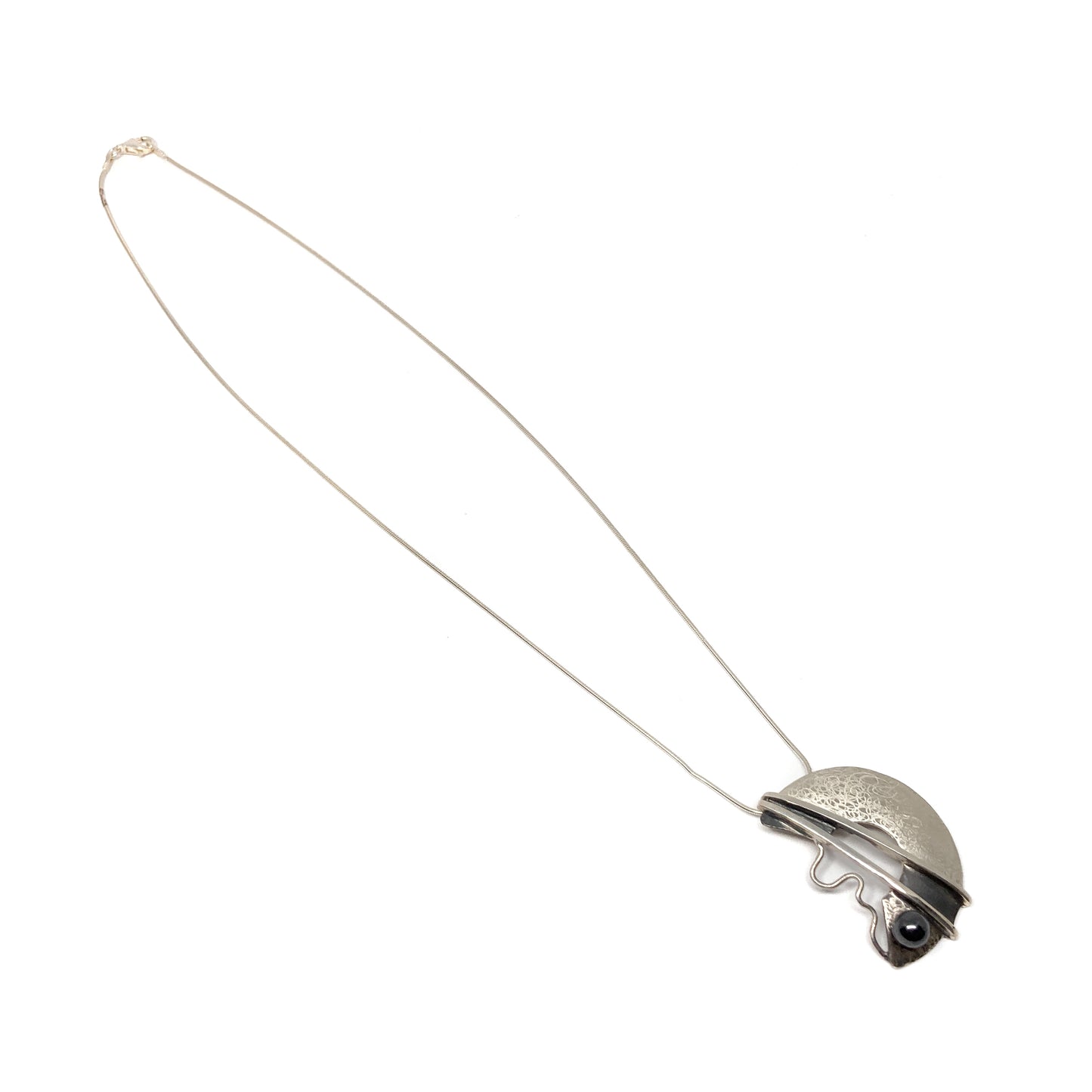 A silver pendant necklace with a semi-circular design, featuring a black bead and abstract lines. The pendant hangs from a delicate silver chain.