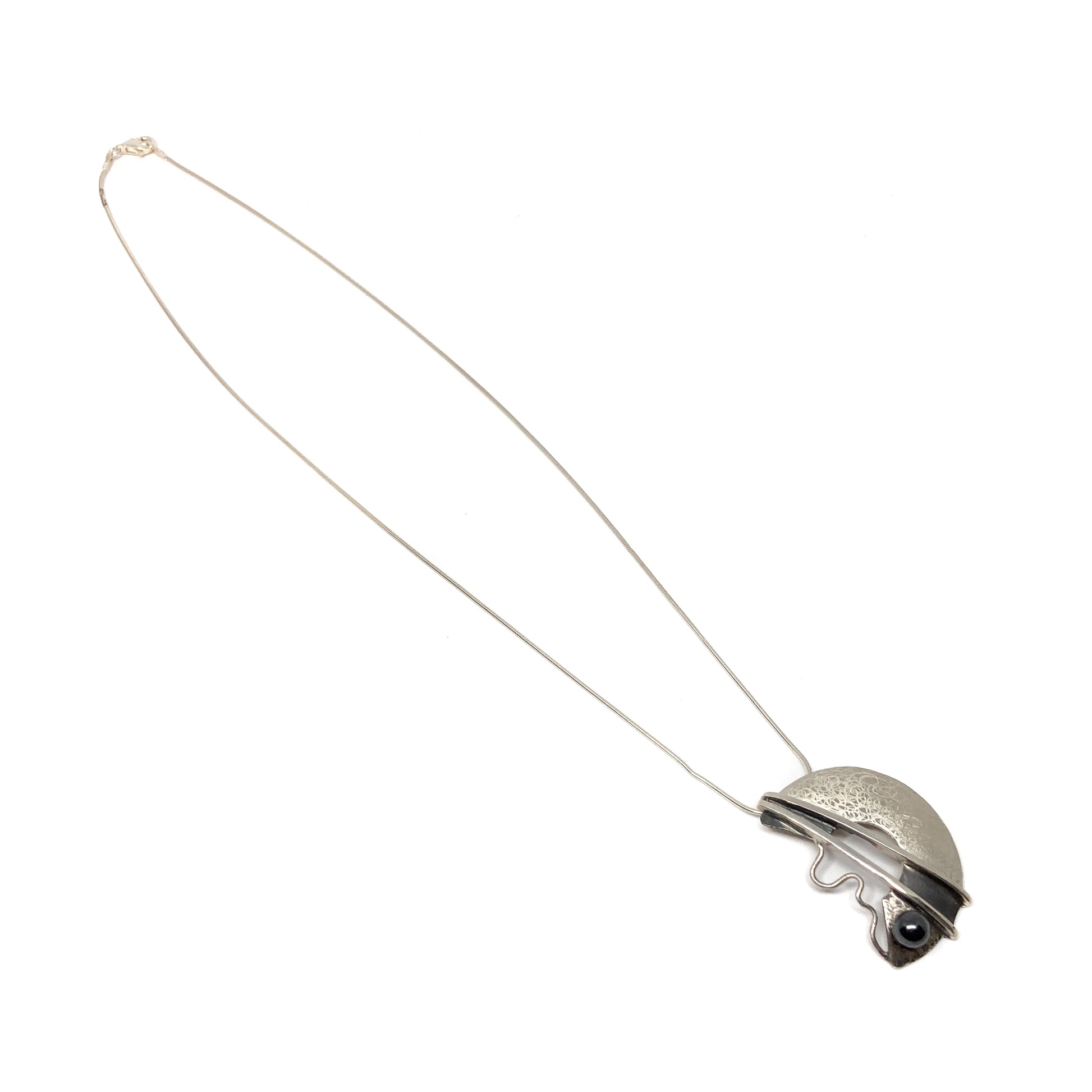 A silver pendant necklace with a semi-circular design, featuring a black bead and abstract lines. The pendant hangs from a delicate silver chain.