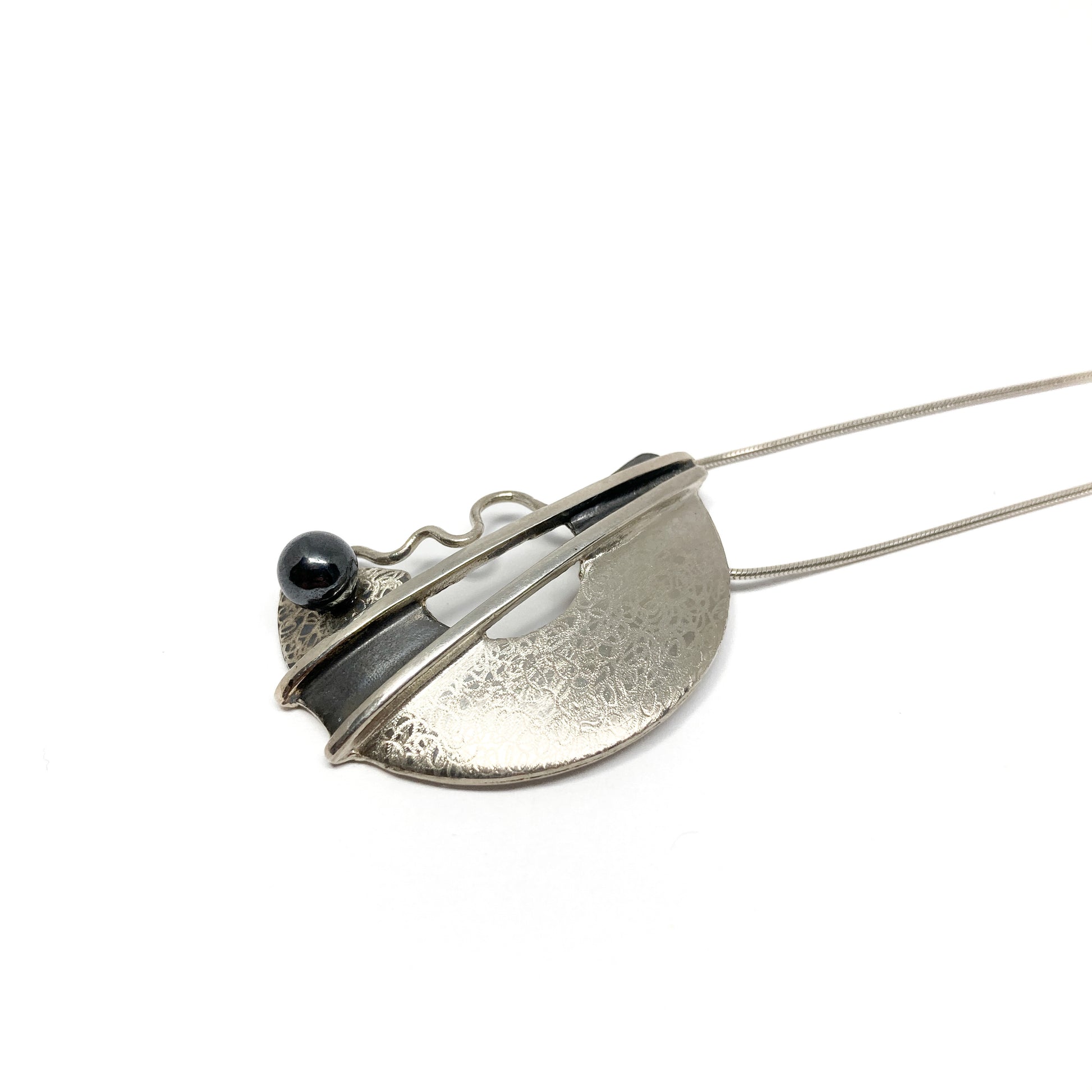 A silver pendant necklace with a semi-circular design, featuring a black bead and abstract lines. The pendant hangs from a delicate silver chain.